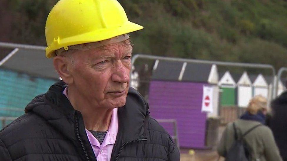 Man wearing yellow hard hat, pink shirt and black coast metal fencing and purple and turquoise beach huts are behind him.