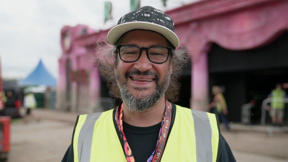 Natty looking direct to camera and smiling. He has on a black and white hat and black rimmed glasses. He is wearing a high-vis vest.