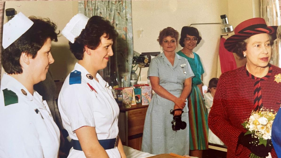 Nurse Mary Donaldson pictured in the hospital wearing a white nursing uniform which has a blue epaulette on the shoulder and a matching blue belt around her waist. She has short brown hair, a white hat and is smiling at the late queen Elizabeth. The late Queen holds flowers on a visit to the ward and wears a red and black checked blazer with a matching hat. 