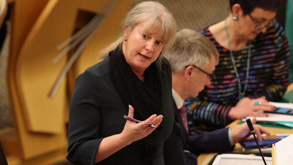 Shona Robison, a woman with blonde hair, speaks in the Scottish Parliament chamber with her right arm, bent at the elbow, held in front of her. She is holding a pen. She is wearing a black top and is visible from the waist up.  