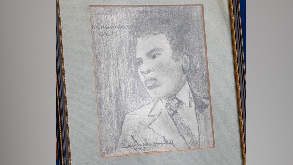A black and white sketch of Muhammad Ali by Eric Morecambe is seen mounted on an easel. It shows a depiction of Ali wearing a suit and tie and is signed by Morecambe