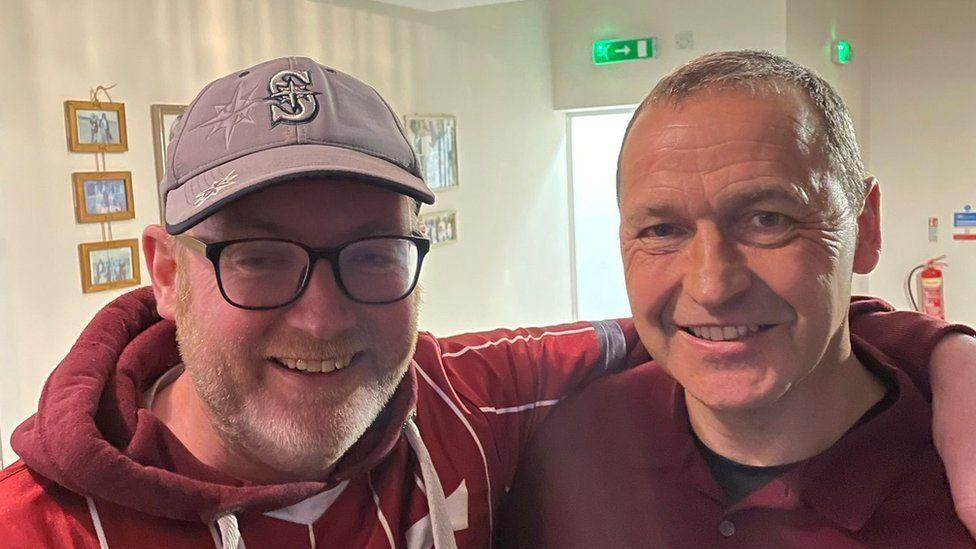 Martin Maloney wearing a cap, glasses and claret hoodie, with his arm around Colin Calderwood. They are both smiling.