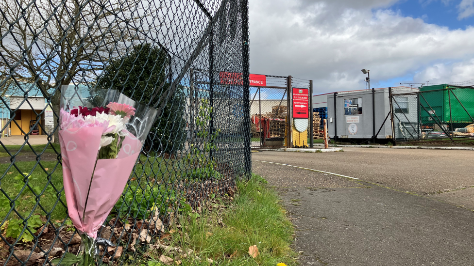 A bouquet of flowers, in pink wrapping, are tied to a metal fence. In the background is a gated road entrance. It is a cloudy day.