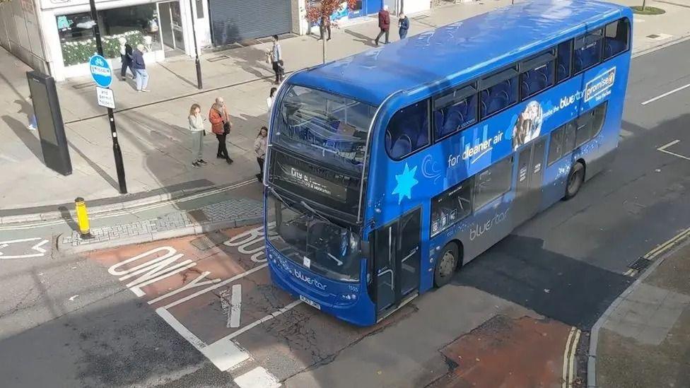 A upper view of a blue Bluestar branded bus in a bus lane with pedestrians passing on