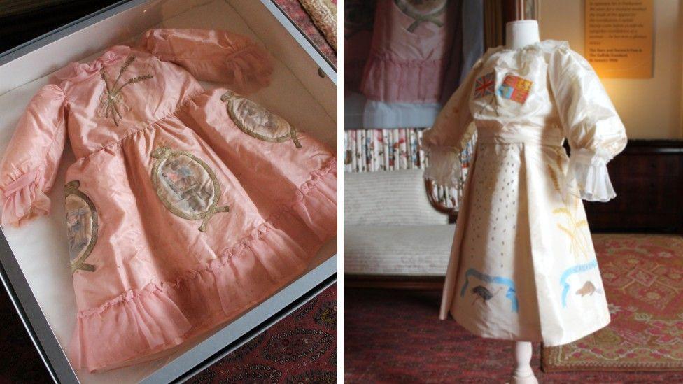 On the left is a small pink silk dress which has wheat stems on the bodice and other emblems across the skirt of the dress. On the right is a white dress displayed on a mannequin which has flags on it and other emblems and ribbons. 