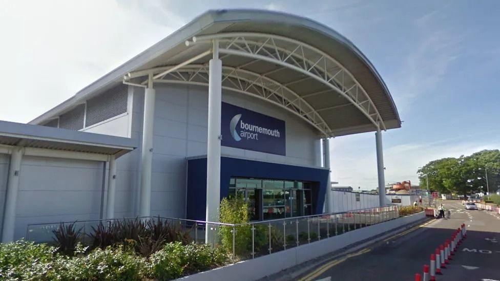 Google Street View of the entrance to Bournemouth airport terminal building. It is a large building clad with white panels and with a large arched portico sticking out over the footpath below. The Bournemouth Airport sign above the glass doors is Navy blue with white writing. The road in front has cones down the middle and a man wearing hi-viz can be seen in the distance.
