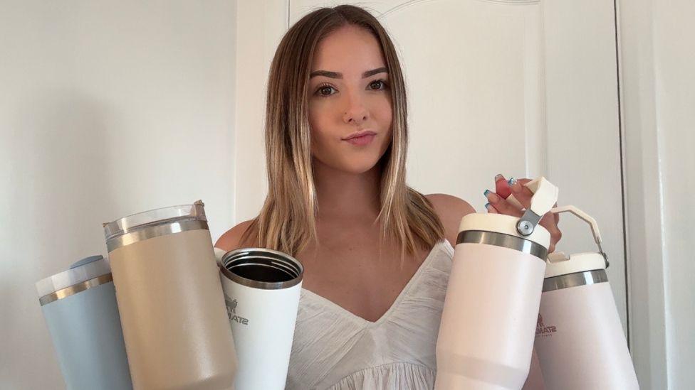 A young woman with light brown hair wearing a white vest holding five Stanley cups