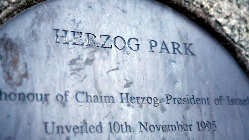 A metal plaque, the text is barely visible but the words Herzog Park are spelled out, also mention of honour of Chaim Herzog, President of Israel, unveiled 10th November 1995