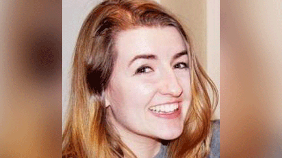 Zoe is smiling at the camera. She has strawberry blonde shoulder-length hair and is wearing a grey top