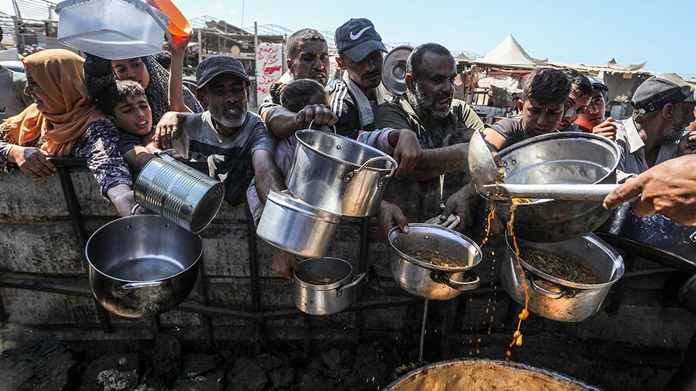 Palestinians, including children, living in the al-Mawasi area wait to receive food distributed by aid groups in Khan Yunis. Men, women and children in drab clothing are crowding around holding out pans and tins as a ladle pours some orange coloured food into one.