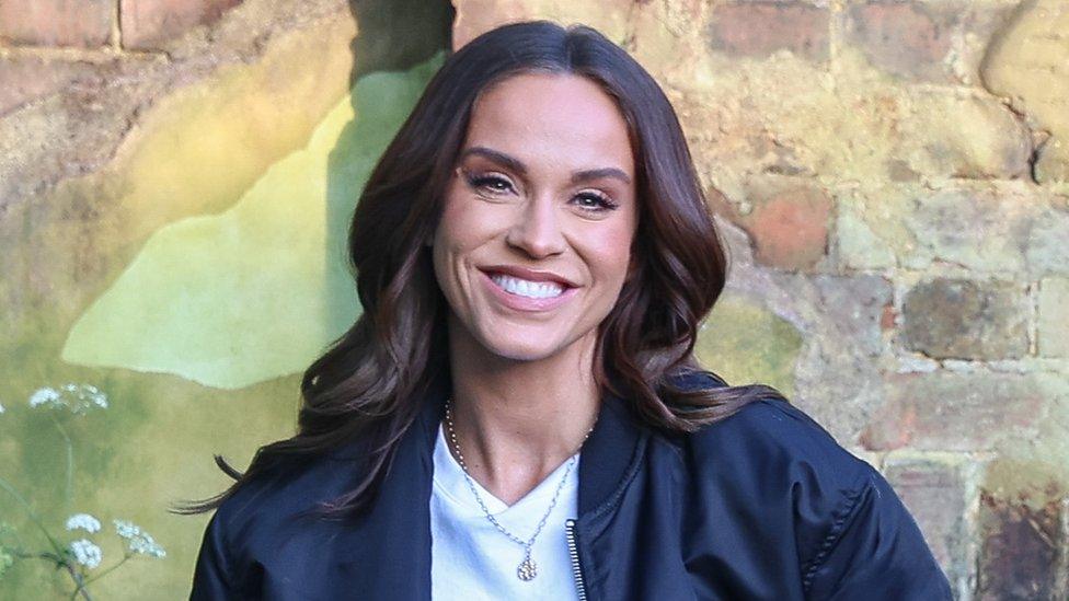 Vicky Pattison pictured in May 2023. Vicky is a 36-year-old white woman with long brown hair and brown eyes. She wears a black bomber jacket over a white T-shirt and a gold chain. She smiles at the camera and is pictured outside against a brick wall