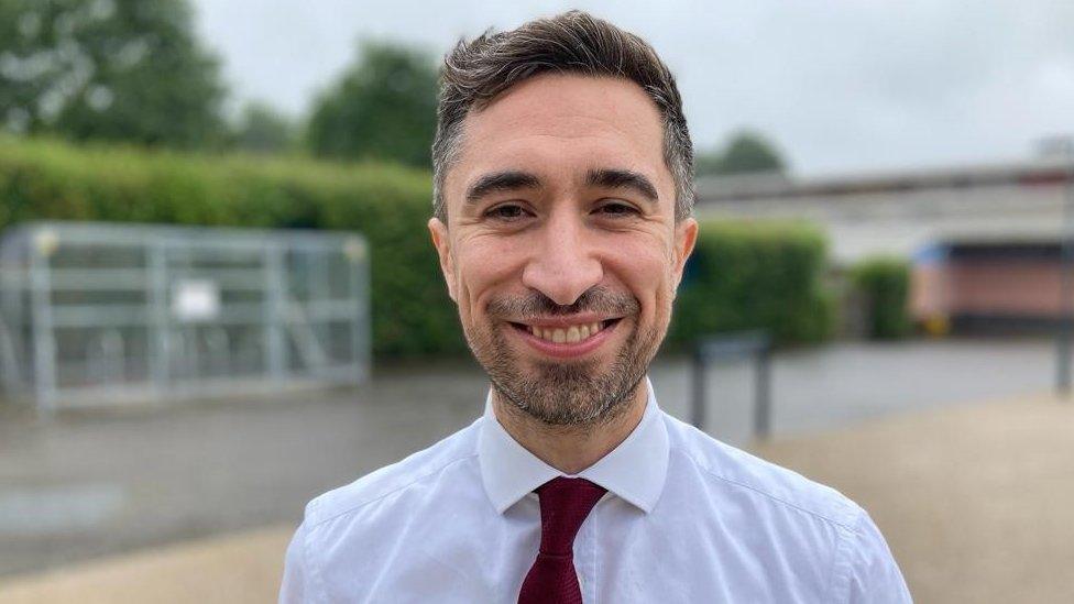 Damian Egan smiles, wearing a white shirt and red tie