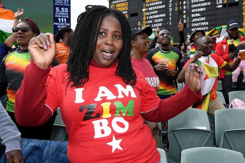 A woman in the stands waves her hands. She is wearing a T-shirt that says 'I am Zimbo'.