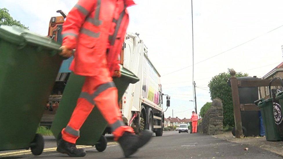 Bin worker pulling along wheelie bins on a pavement, with a bin lorry in the background