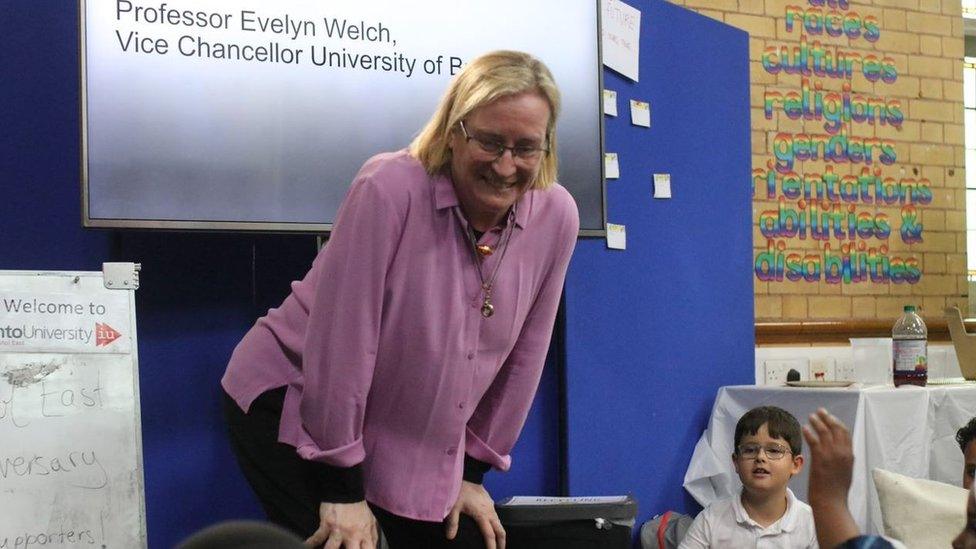 A blonde woman in a lilac top smiling down at children which are sitting around her