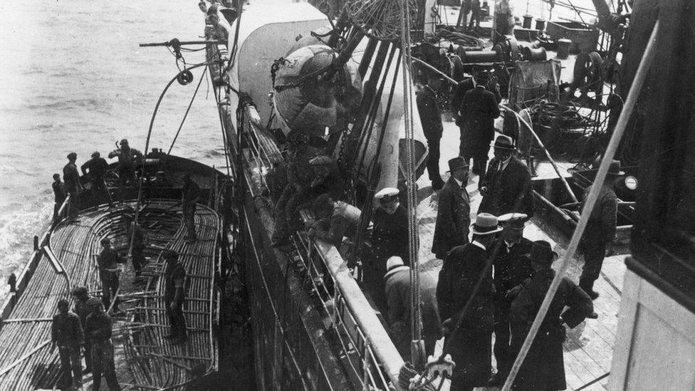 Groups of men on the deck of a ship which laying telegraph cable at sea, with the image showing men looking over the side of the ship at a smaller vessel carrying the cable, circa 1900