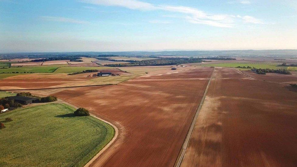 An aerial view of a brown farm field with a concrete line down the middle indicating the location of the former Tarrant Rushton airfield