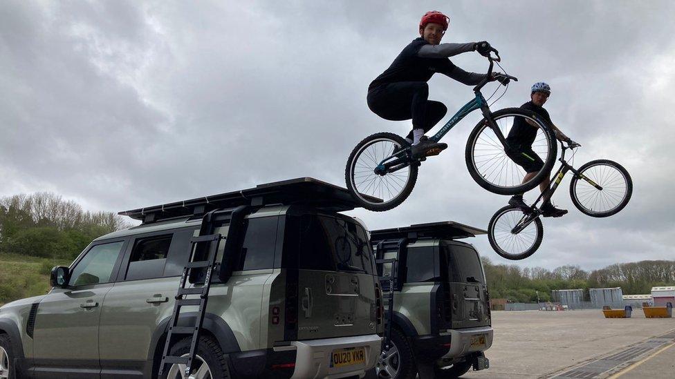 Ashley and Danny Butler on bikes, jumping off the roof of two Land Rovers