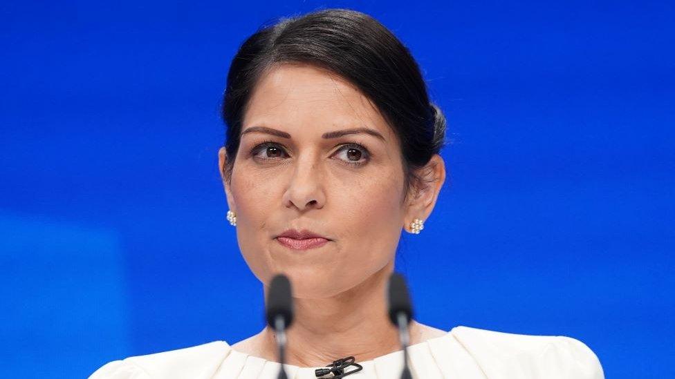 Priti Patel standing at a microphone.