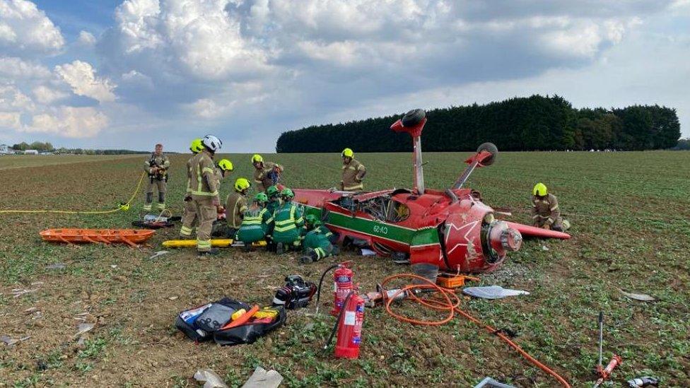 A red light aircraft upside down. Wheels are sticking up and it has been surrounded by emergency services including paramedics and firefighters