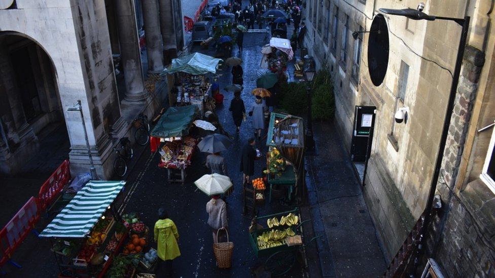 Image of Frog Lane. Cast members can be seen walking down the street, holding umbrellas. The set is a London market in the 1970s.