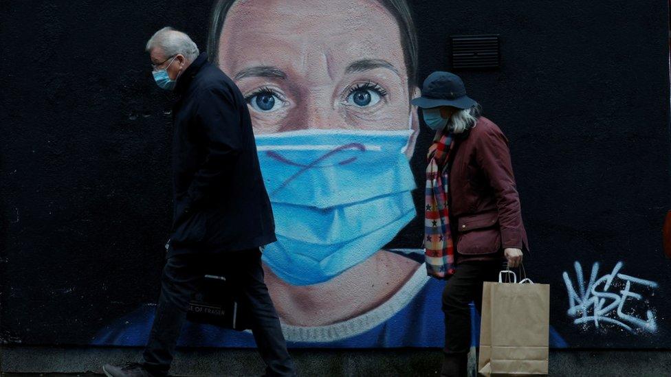 Two people walk past wall art of health worker wearing mask