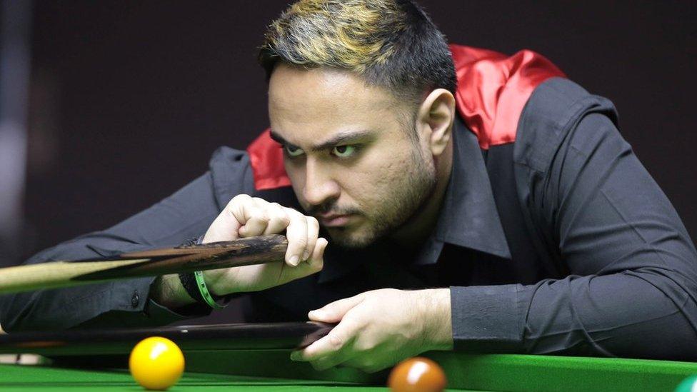 An Asian snooker player wearing a smart black shirt and waistcoat with a velvety red back leans over the green baize of a snooker table. He's concentrating hard as he holds a cue in one hand and a rest with his other. A yellow, brown and green ball can be seen on the table.