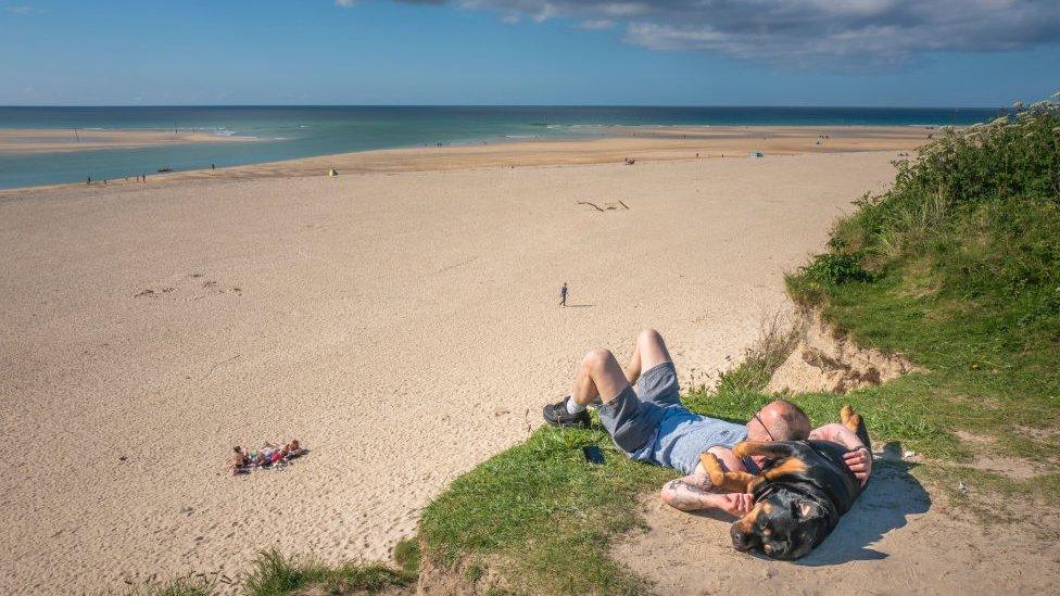 A man sunbathing with his dog in sand dunes looking out at the beach and sea