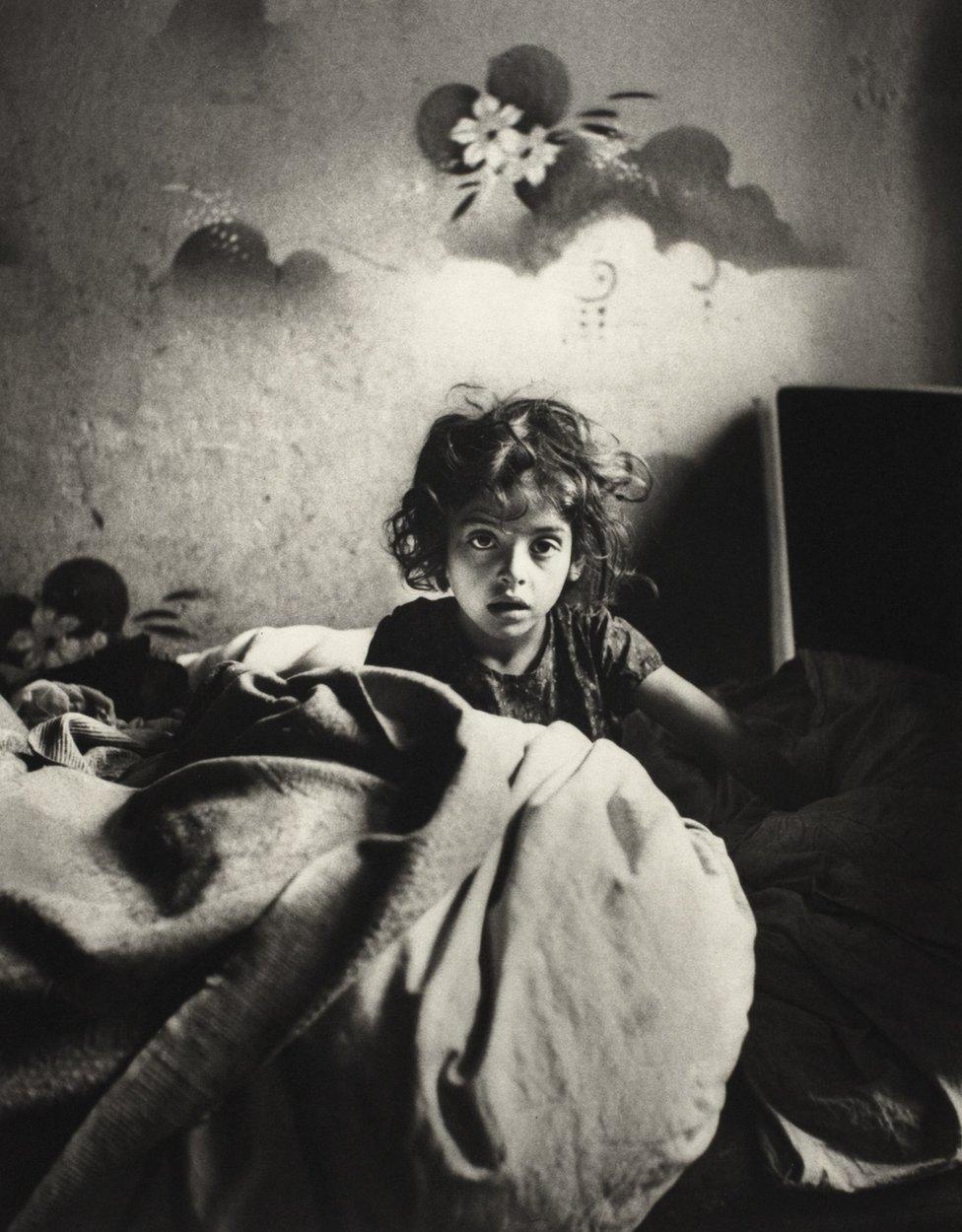 Sara, sitting in bed in a basement dwelling, with stencilled flowers above her head. Warsaw, Poland, c. 1935–37.