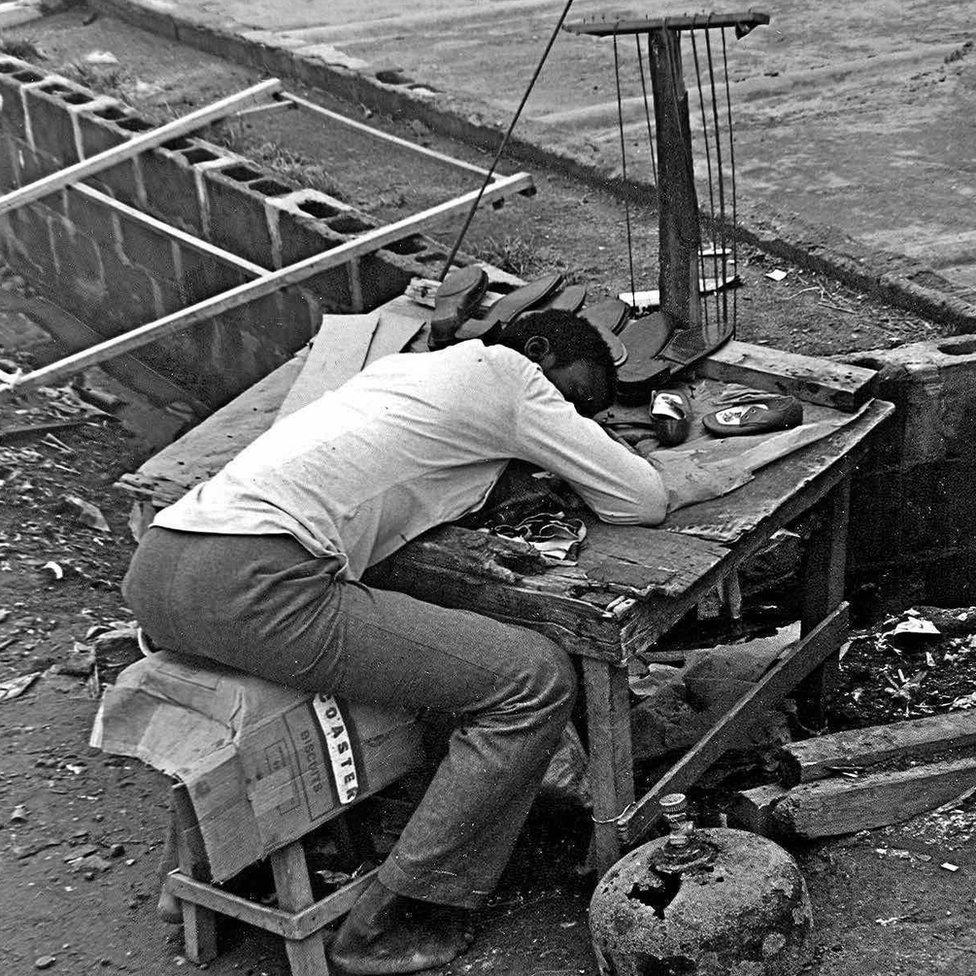 A photo by Sunmi Smart-Cole entitled: "Wake Up, Nigeria" - 1984, a man a cobbler asleep on his work bench