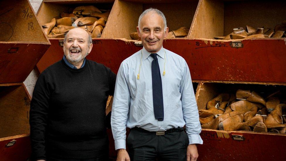 Two men, one wearing a tie and the other a blue sweater, stand in front of containers of part-built shoes