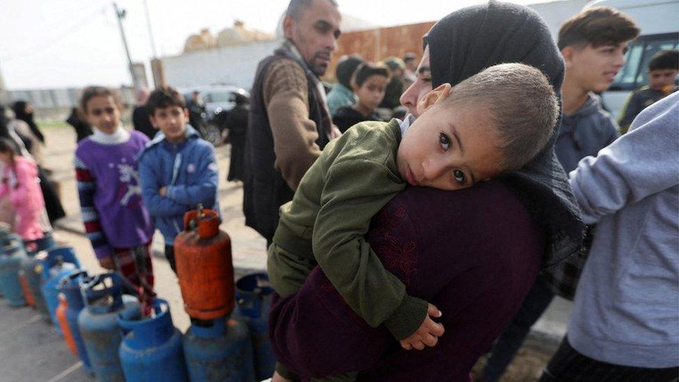 A woman carries a child as Palestinians gather to fill liquid gas cylinders, during a temporary truce between Hamas and Israel, in Rafah in the southern Gaza Strip