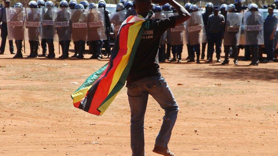 A man wearing a Zimbabwean flag salutes riot police during a protest in Harare, Zimbabwe, on Friday 26 August 2016