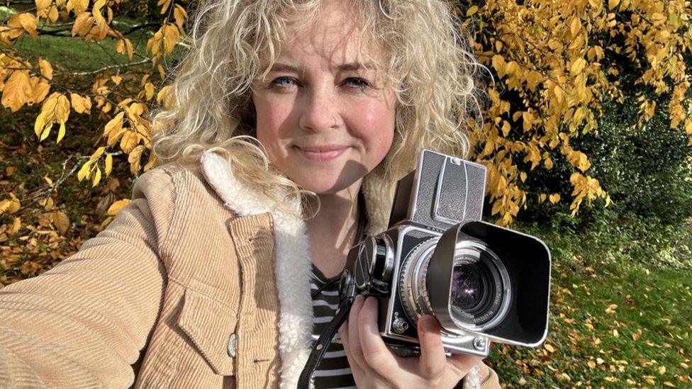 Cíara Hillyer holding a camera and smiling in front of a tree with orange leaves