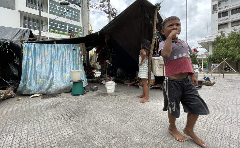 An Emberá chid stand next to a makeshift shelter in Montería