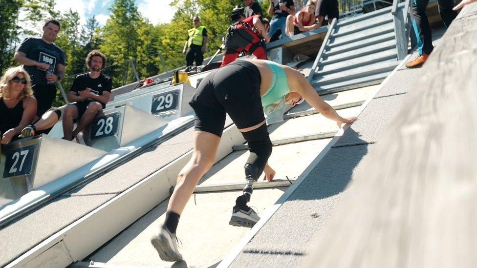 Milly Pickles near the finish line of the Red Bull 400 race. Milly is crawling up the steep incline, which is paved. She wears black shorts and a green-blue sports top. Her prosthetic leg is visible on her right and behind her spectators cheer her on.