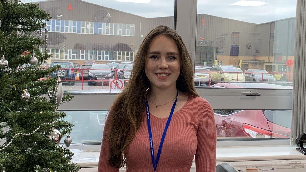 Abi smiling with a factory behind her and next to a christmas tree