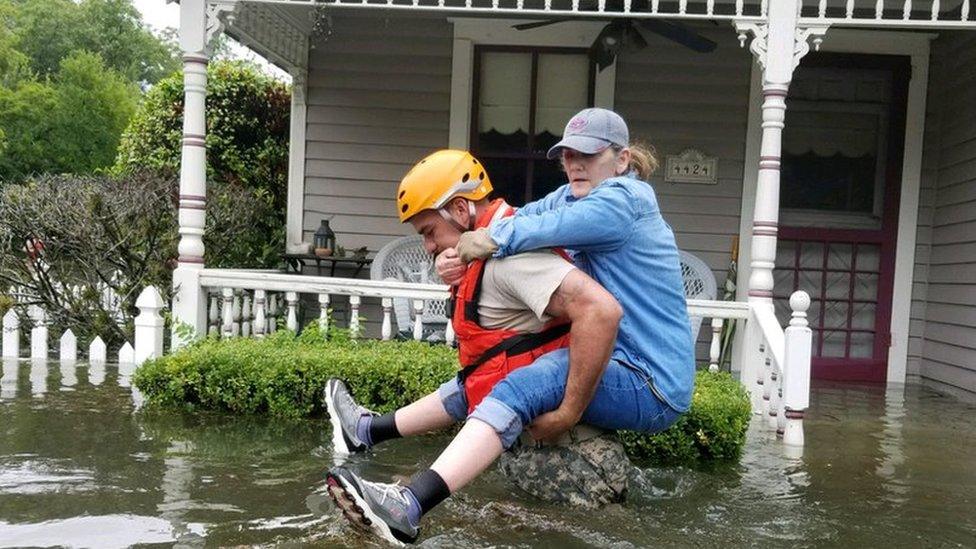 A soldier, waist-deep in water, carried a woman on his back past the front of a house porch