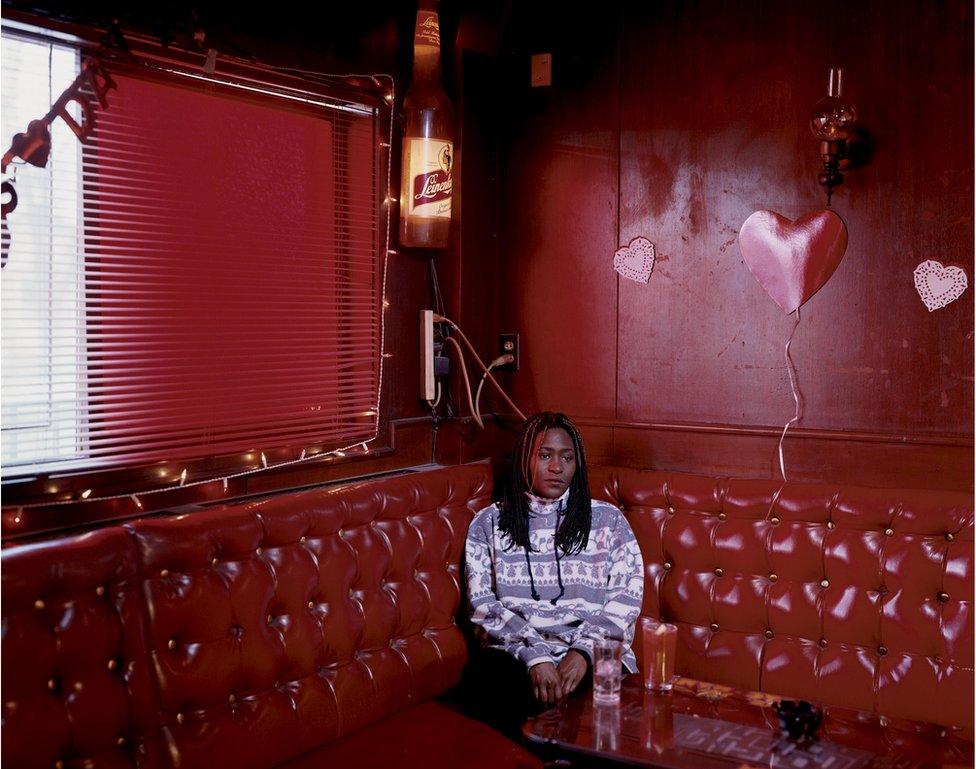 Kym sits in the corner of a nightclub with red walls and sofas