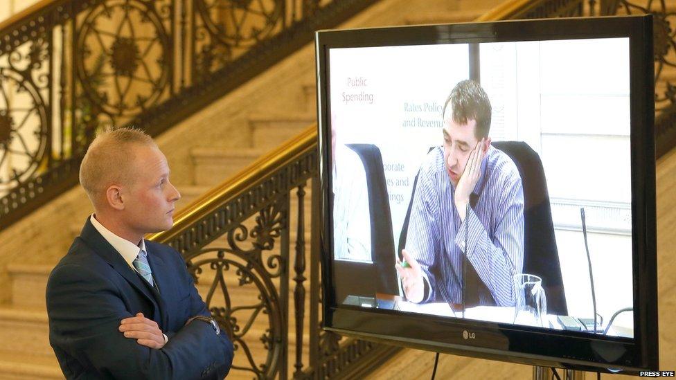 Jamie Bryson looks at TV screen showing Daithí McKay in Stormont, file pic from September 2015