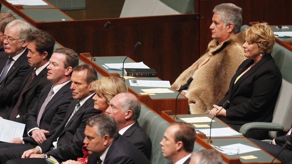 Ken Wyatt, wearing a traditional kangaroo skin coat, sits in parliament with other MPs