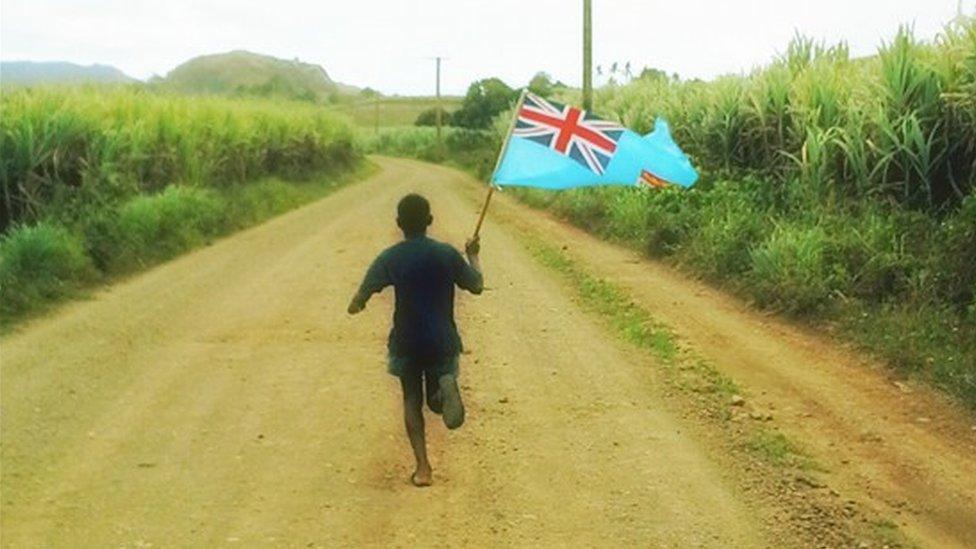 A Fiji boy runs down a dirt road, cheering on the rugby sevens team