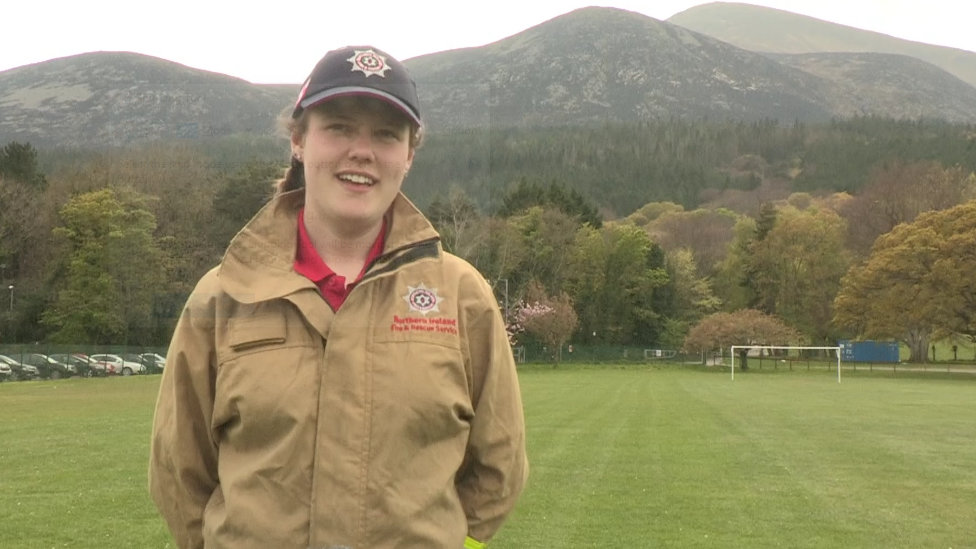 Hayley Agnew dressed in a fire service jacket, standing in front of the Mourne Mountains in the daytime, smiling with a fire service baseball cap on