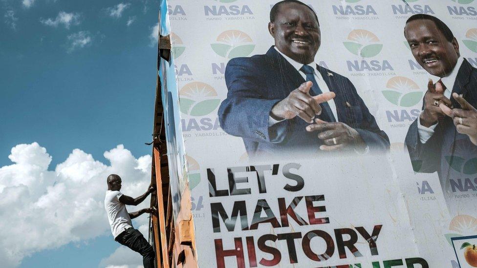 A man climbs the scaffolding of a National Super Alliance electoral poster, set against a bright blue sky. The poster, featuring Raila Odinga, reads: "Let's make history together"