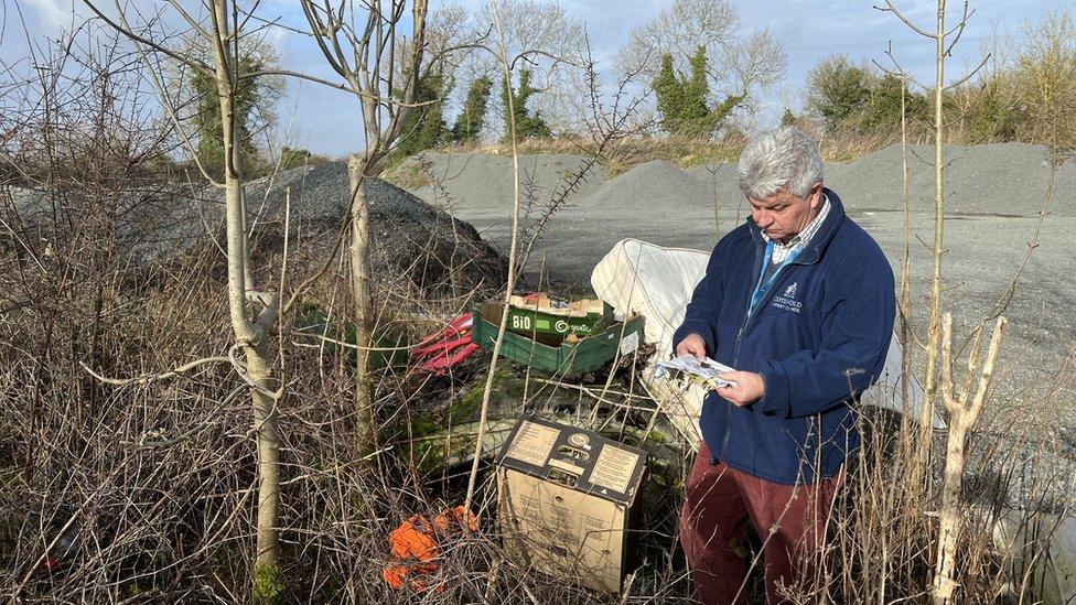 A man in a council branded blue fleece holding a note stands next to dumped waste, including a mattress and boxes