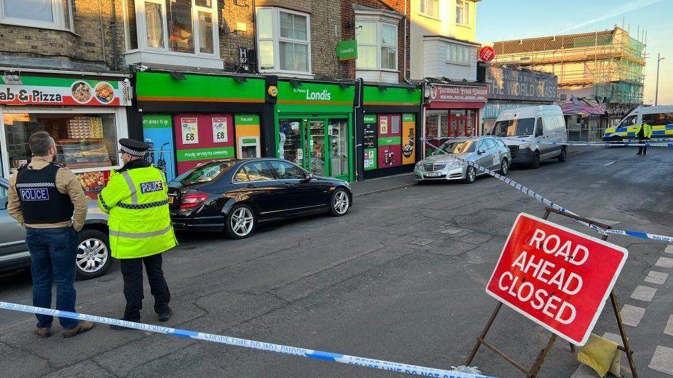 Two police officers facing away from the camera looking at a cordon. There is a red road closed sign next to him at a road junction which is also surrounded by police tape. There is a row of shops also within the cordon as well as several cars parked on the pavement. 