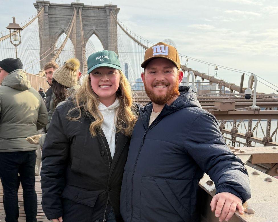 A woman and a man stand on a bridge in New York, smiling at the camera. They are both wearing coats and baseball caps