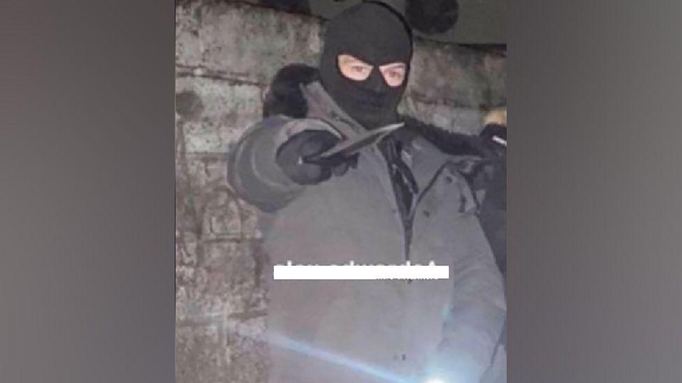 Alex Hutton wears a balaclava and poses with a machete