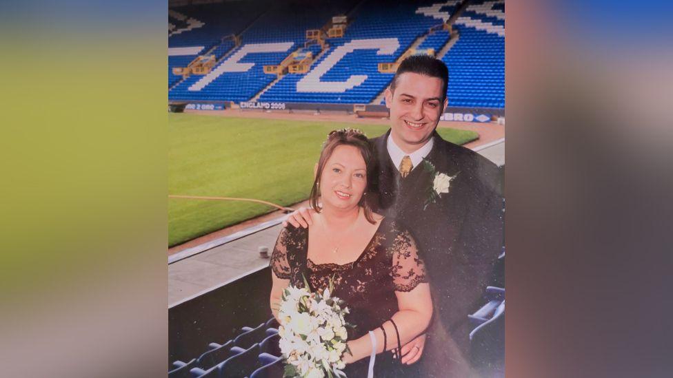 Del and Aly Thornton are standing in the main stand at Goodison Park on their wedding day. Aly is wearing a black lace dress and holding a bouquet of white flowers while Del is wearing a suit.