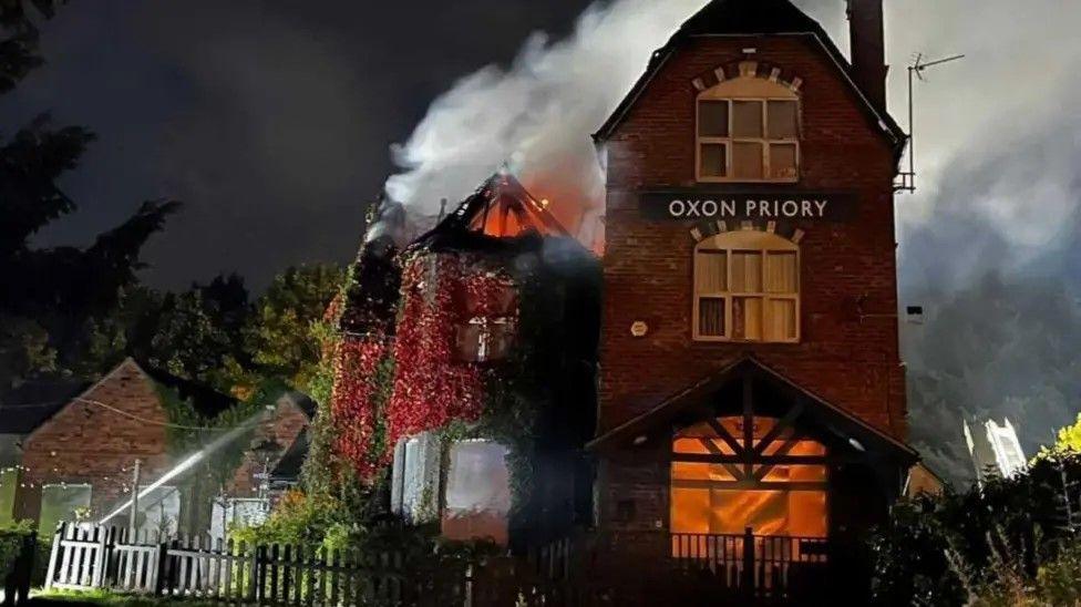 A burning three-storey building with sharply sloped roofs. There are bushes and trees either side of the building and a jet of water can be seen aimed at the second storey.